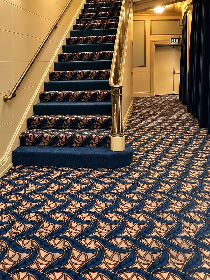 Staircase with brass handrail and blue patterned carpet runner leading into a hallway with matching patterned carpet